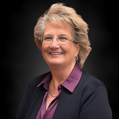 Smiling older woman with short, light hair and glasses, wearing a purple blouse and dark blazer, posing against a plain black background.