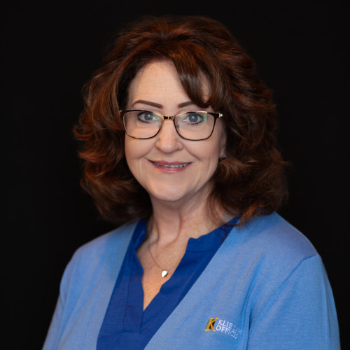 A woman with curly brown hair, wearing glasses and a light blue cardigan over a blue shirt, smiles at the camera against a plain black background.
