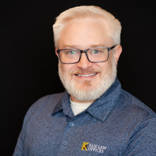 A smiling man with short white hair, glasses, and a beard wears a blue collared shirt with a "Kile Law Offices" logo, posing against a plain black background.