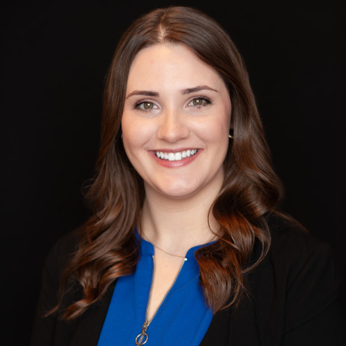 A woman with long brown hair, wearing a blue top and a black blazer, smiles at the camera against a plain black background.
