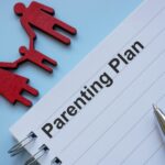 A notebook page titled "Parenting Plan" with a blue pen beside it. Above the page are red cutouts of two adults holding hands with a child, symbolizing a family—ideal for outlining custody arrangements for unmarried parents in West Virginia.
