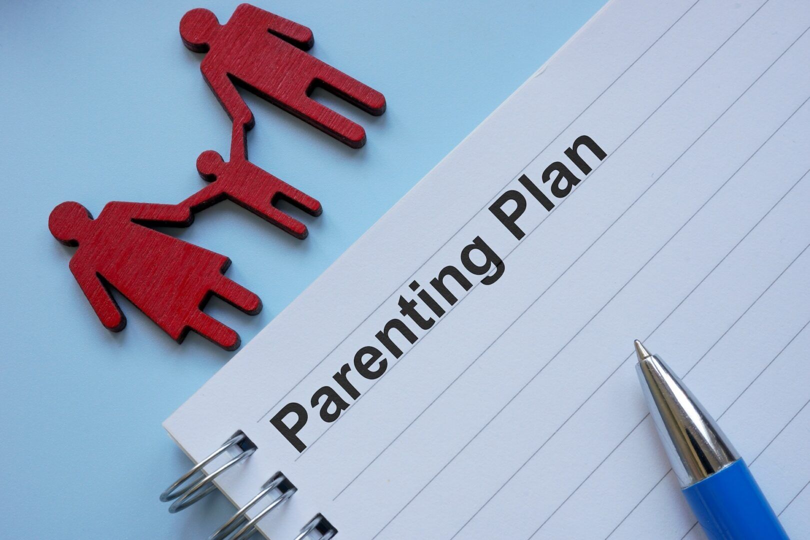 A notebook page titled "Parenting Plan" with a blue pen beside it. Above the page are red cutouts of two adults holding hands with a child, symbolizing a family—ideal for outlining custody arrangements for unmarried parents in West Virginia.