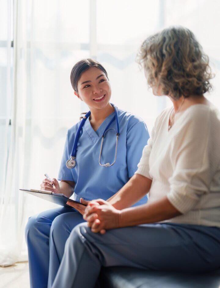 A healthcare professional in blue scrubs sits with a clipboard, smiling and talking to an older woman with curly hair, both seated indoors in a well-lit room.