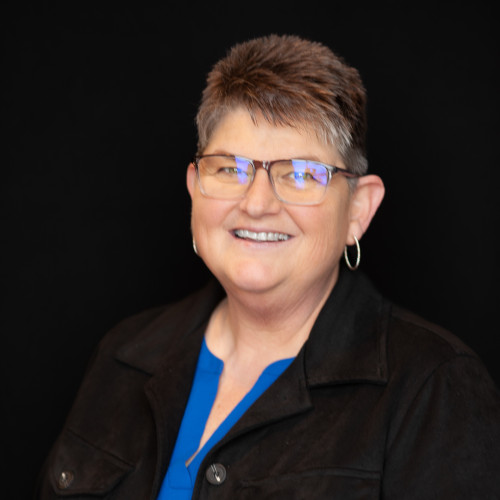 A smiling woman with short gray hair, glasses, and hoop earrings, wearing a black jacket over a blue top, posing in front of a plain black background.