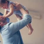 A man lifts a smiling toddler indoors, sharing a playful moment that highlights the joy of fatherhood. The child touches his face as they laugh together—a touching scene reflecting fathers' rights and bonding in West Virginia custody situations.