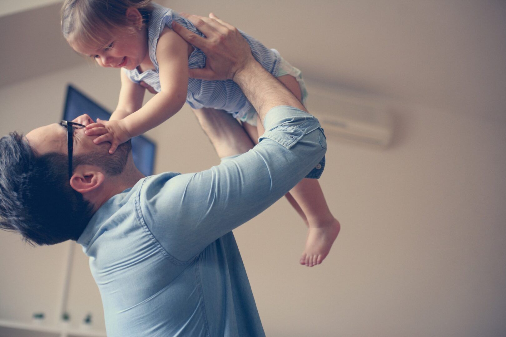A man lifts a smiling toddler indoors, sharing a playful moment that highlights the joy of fatherhood. The child touches his face as they laugh together—a touching scene reflecting fathers' rights and bonding in West Virginia custody situations.