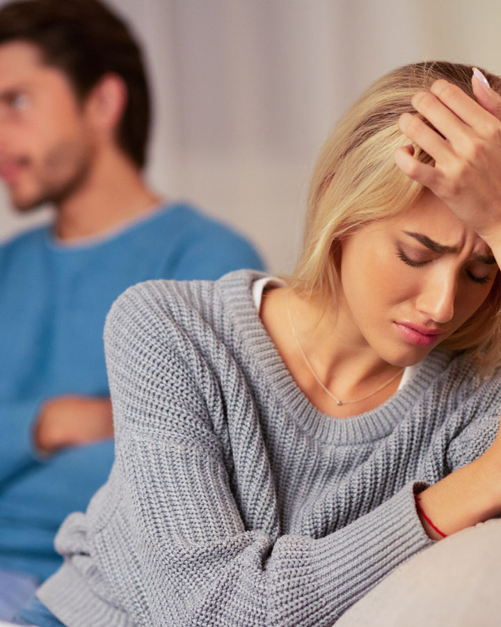 A woman sits on a couch looking upset with her hand on her forehead, while a man in the background sits turned away, both appearing distressed after an argument.