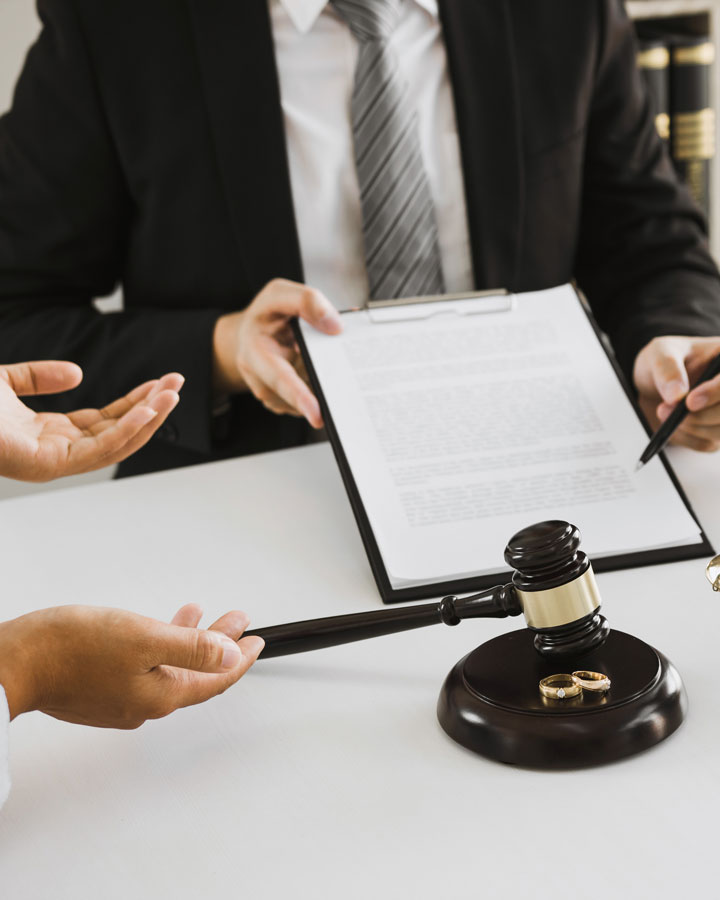 A person in a suit holds a clipboard with a document while another points to it. On the table are a gavel and two wedding rings, representing a legal or divorce discussion.