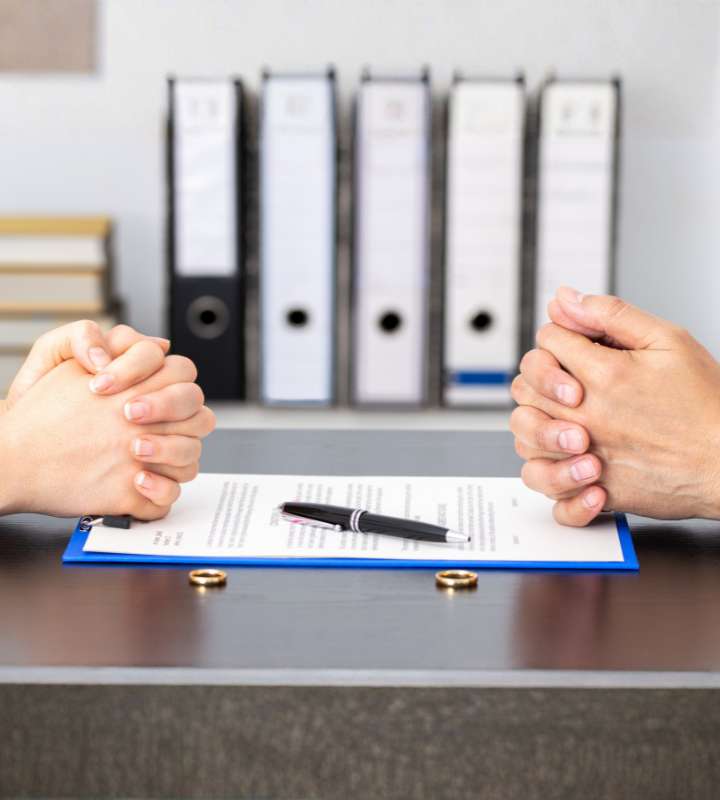 Two people sit across a desk with hands clasped, facing each other over a document, pen, and two wedding rings, suggesting a divorce or legal agreement. Binders and books are in the background.