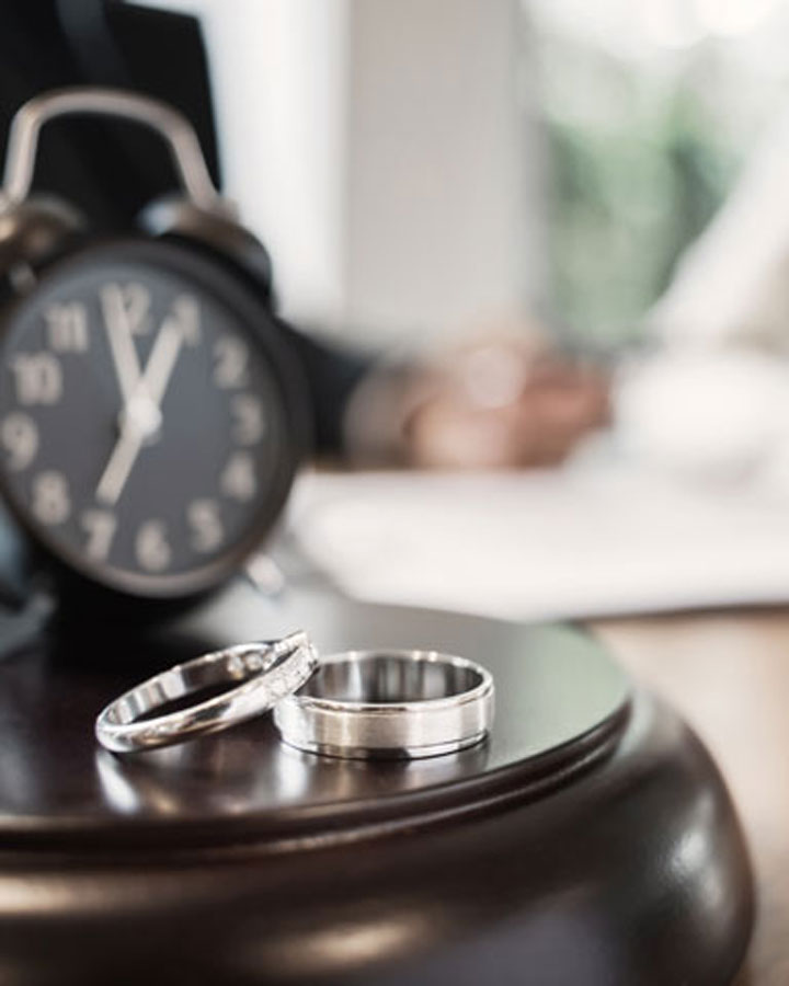 Two silver wedding rings rest on a wooden surface next to a blurred clock, with a person in the background holding paperwork, suggesting themes of marriage, time, or possibly divorce.