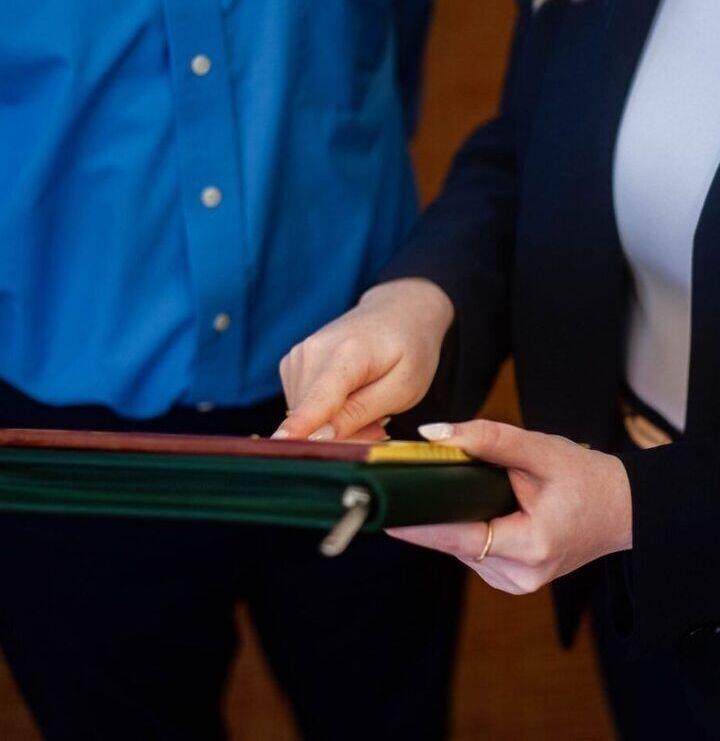 Two people stand together; one in a blue shirt, the other in a dark blazer and white top, pointing at documents inside a green folder. Their faces are not visible.