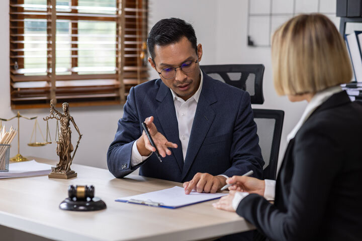 A man in a suit gestures while speaking to a woman across a desk in an office. Legal symbols, including a gavel and Lady Justice statue, are on the desk, suggesting a legal consultation or meeting.