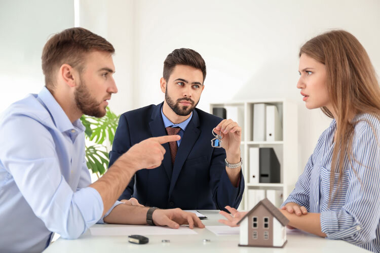 A man and woman argue across a table while a serious-looking man in a suit, holding house keys, sits between them. A small model house is on the table, suggesting a disagreement over property or real estate.