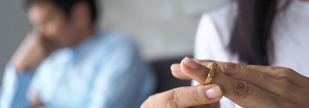 A person removes a wedding ring from their finger, with another person sitting in the background looking upset or distressed. The focus is on the hand and the ring, symbolizing relationship or marital issues.