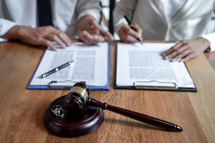 Two people sit at a table, reviewing and signing documents on clipboards. A gavel and two wedding rings are placed on the table in the foreground, suggesting a legal or divorce proceeding.