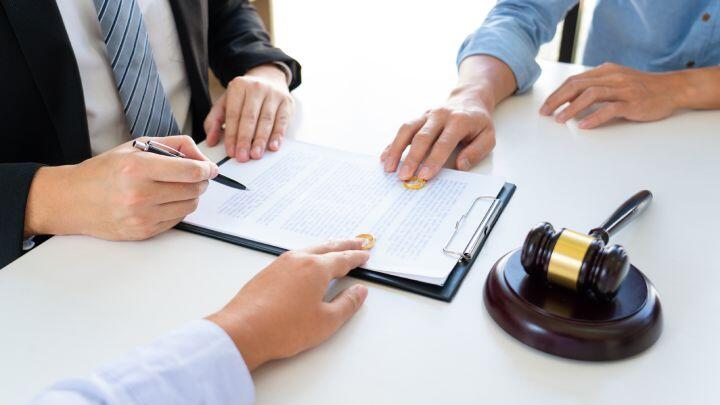 Three people sit at a table reviewing a document on a clipboard. One points with a pen, another gestures at the paper, and a gavel rests on the table, suggesting a legal or contract discussion.