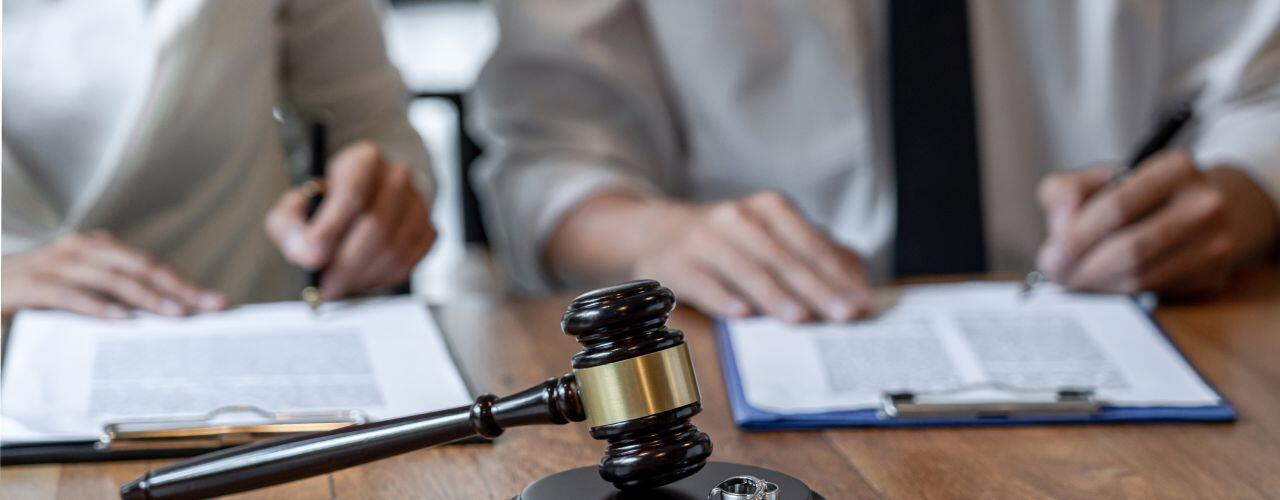 A judge's gavel and two wedding rings on a table, with two people in business attire sitting in the background, signing documents on clipboards.