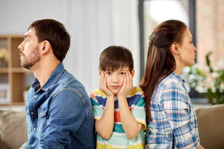 A sad boy sits between a man and a woman, both facing away from each other with serious expressions. The boy rests his face in his hands, looking concerned. They are indoors on a couch.