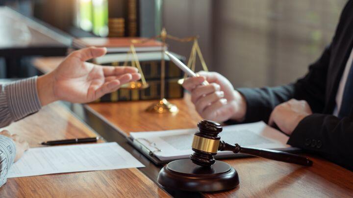 Two people sit at a desk with legal documents, a gavel, and scales of justice. One person gestures with their hand while the other holds a pen, suggesting a legal discussion or consultation.