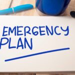 An open notebook with the words "EMERGENCY PLAN" written in blue marker—perfect for outlining steps for emergency custody in West Virginia—sits beside a blue pen and cup on a wooden desk.
