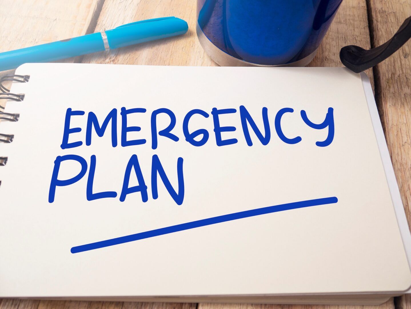 An open notebook with the words "EMERGENCY PLAN" written in blue marker—perfect for outlining steps for emergency custody in West Virginia—sits beside a blue pen and cup on a wooden desk.