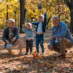 Two adults, possibly grandparents, and two young children play together in a park filled with autumn leaves. The adults crouch and smile, while the children stand tossing leaves joyfully. Tall trees with yellow leaves are in the background.