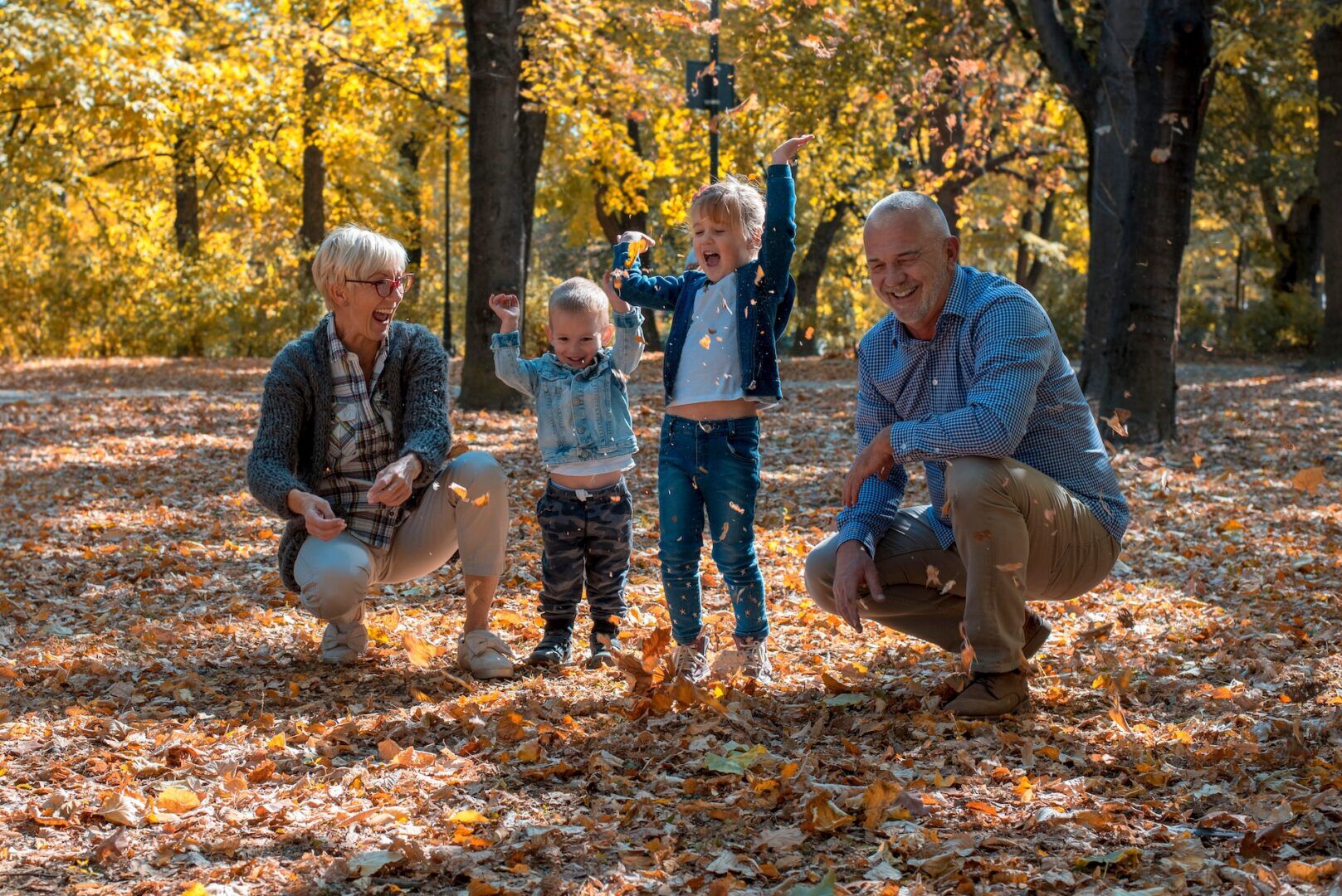 Two adults, possibly grandparents, and two young children play together in a park filled with autumn leaves. The adults crouch and smile, while the children stand tossing leaves joyfully. Tall trees with yellow leaves are in the background.