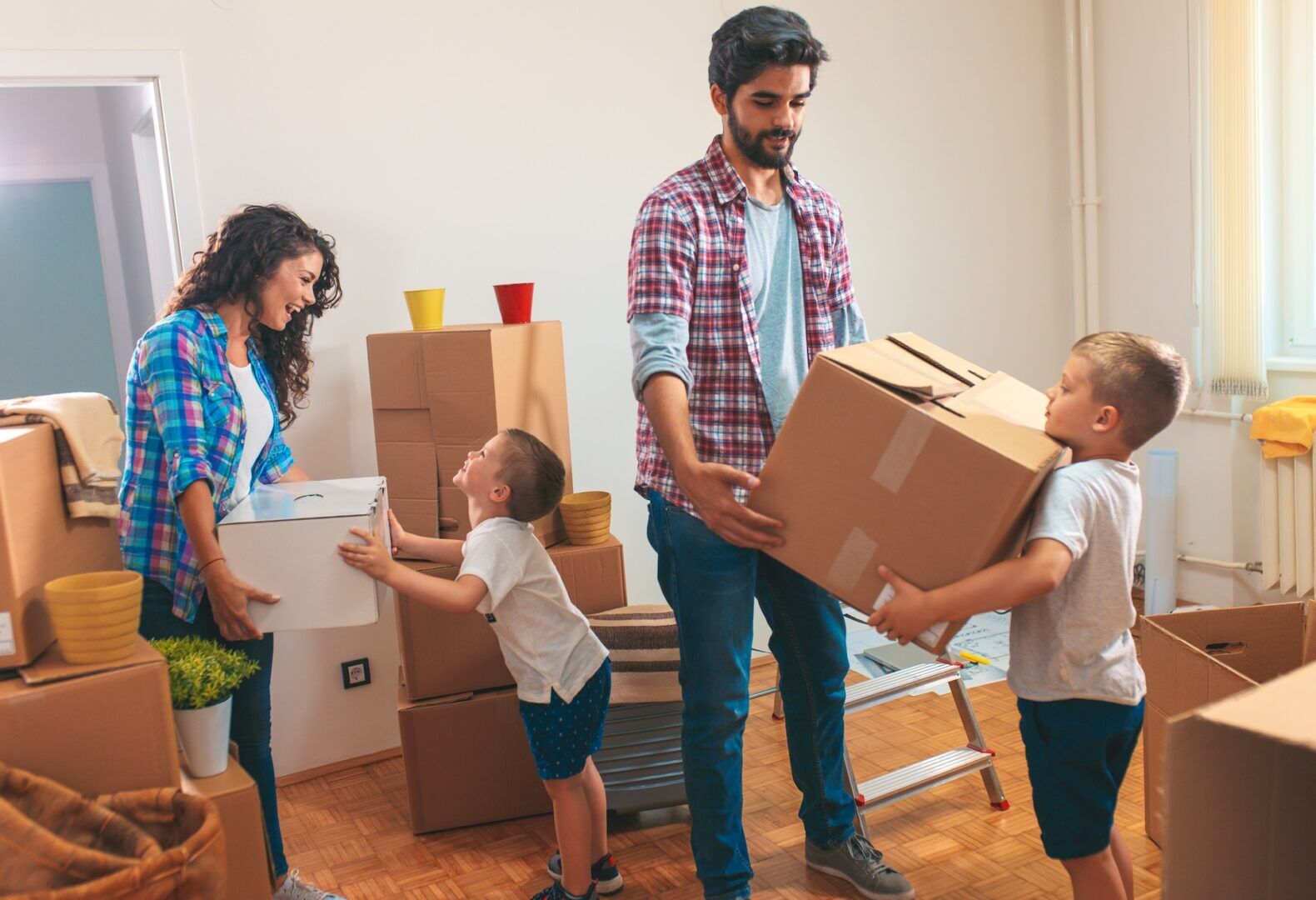A family of four, including two young children, is packing or unpacking moving boxes in a bright room with wooden floors and scattered boxes—perhaps during an interstate custody move between West Virginia and Ohio. The adults smile as the children help carry a box.