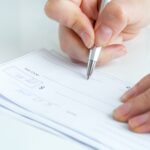 A close-up of a hand holding a pen and writing on a check—possibly related to Ohio spousal support alimony factors—with two more checks underneath on a white surface.