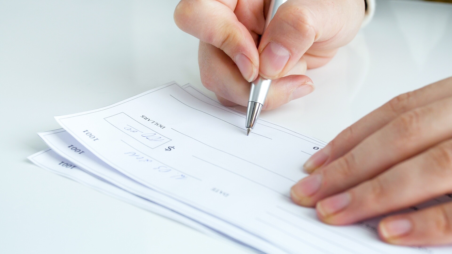 A close-up of a hand holding a pen and writing on a check—possibly related to Ohio spousal support alimony factors—with two more checks underneath on a white surface.