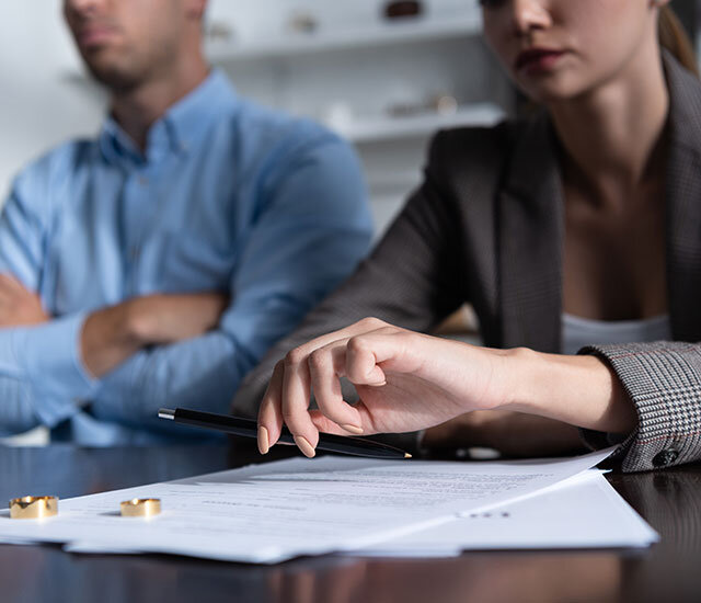 Two people sit at a table with divorce papers and two wedding rings in front of them. One person holds a pen, ready to sign, while the other sits with arms crossed. Both have serious expressions.