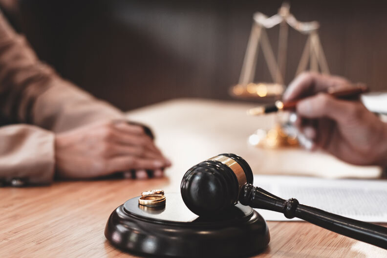 A judge’s gavel and two wedding rings rest on a table, with two people sitting across from each other, one holding a pen, and a legal document and scales of justice in the background.