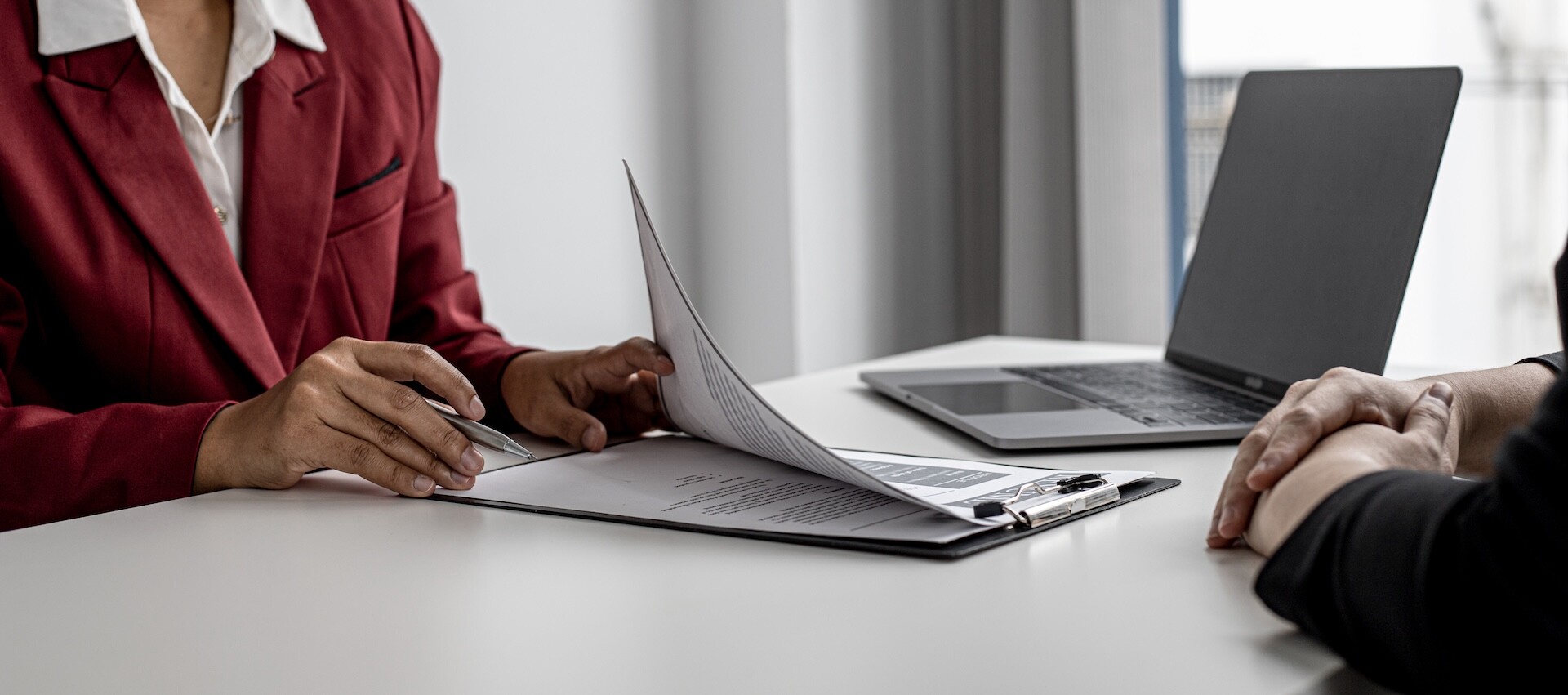 Two people sit across a desk from each other; one holds a pen and points to a document on a clipboard, while the other has hands clasped. A laptop is on the table near the window.