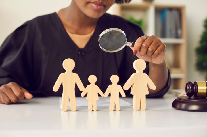 A judge examines wooden cutouts of a family with a magnifying glass at a desk, with a gavel visible nearby. A judge examines wooden cutouts of a family with a magnifying glass at a desk, with a gavel visible nearby.
