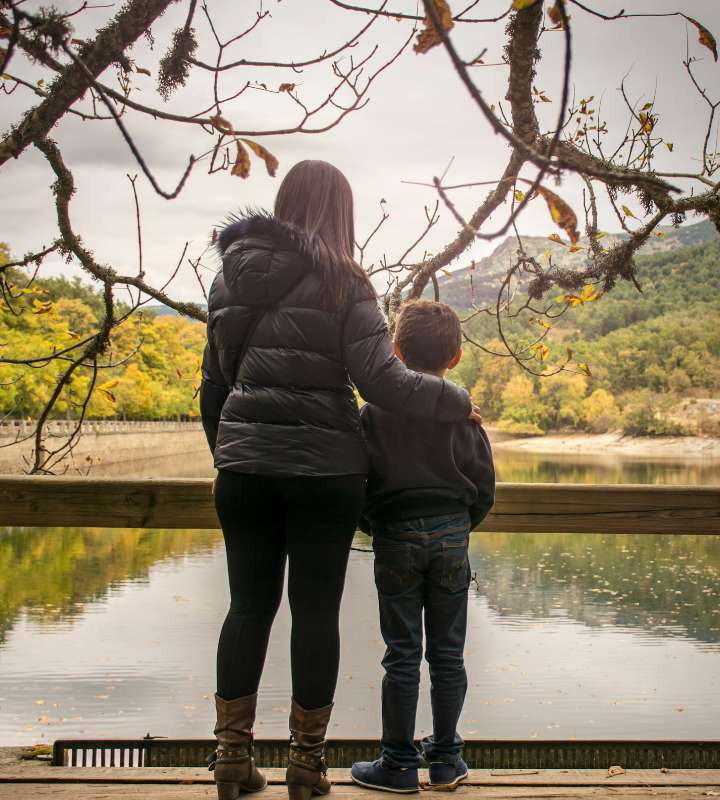 A woman and a child stand side by side on a wooden platform by a lake, looking at the autumn-colored trees and hills across the water. The woman has her arm around the child, and tree branches frame the scene above them. A woman and a child stand side by side on a wooden platform by a lake, looking at the autumn-colored trees and hills across the water. The woman has her arm around the child, and tree branches frame the scene above them.