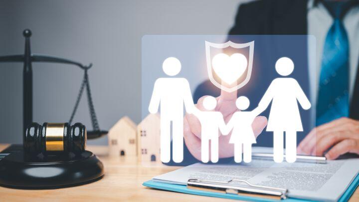 A person in a suit sits at a desk with legal documents, a gavel, and house models. Overlaid is an icon of a family under a shield, symbolizing legal protection or family law.