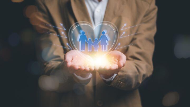 A person in a brown suit holds their hands out, palms up, with a glowing heart-shaped hologram featuring a family icon—two adults and a child. Blue arrows point outward from the heart.