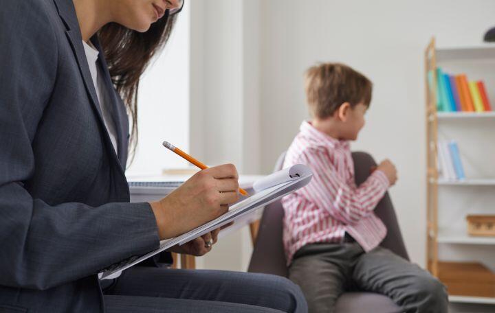 A woman in a suit takes notes on a clipboard while a young boy in a striped shirt sits on a chair, turned away and looking to the side in a room with shelves and books.