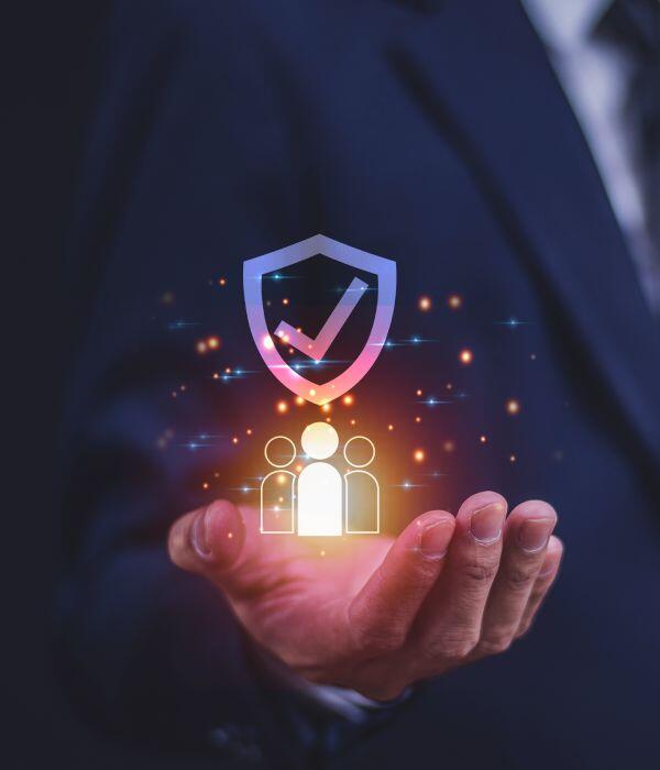 A person in a suit holds out their hand with a glowing hologram of three people and a shield with a checkmark above them, symbolizing security and protection.