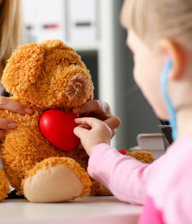 A child holds a toy stethoscope to a red heart on a stuffed teddy bear, while an adult helps support the bear. The scene is playful and nurturing, suggesting a pretend medical checkup.