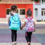 Two young girls with backpacks, holding hands, walk toward the entrance of a colorful school building on a paved walkway.