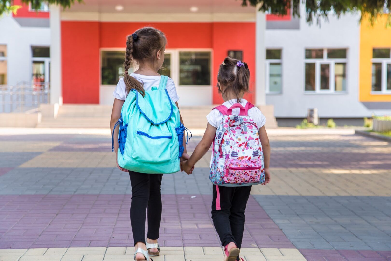 Two young girls with backpacks, holding hands, walk toward the entrance of a colorful school building on a paved walkway.