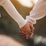 Two people wearing long-sleeved shirts are holding hands outdoors. The image is focused on their clasped hands, with a blurred natural background and warm lighting.