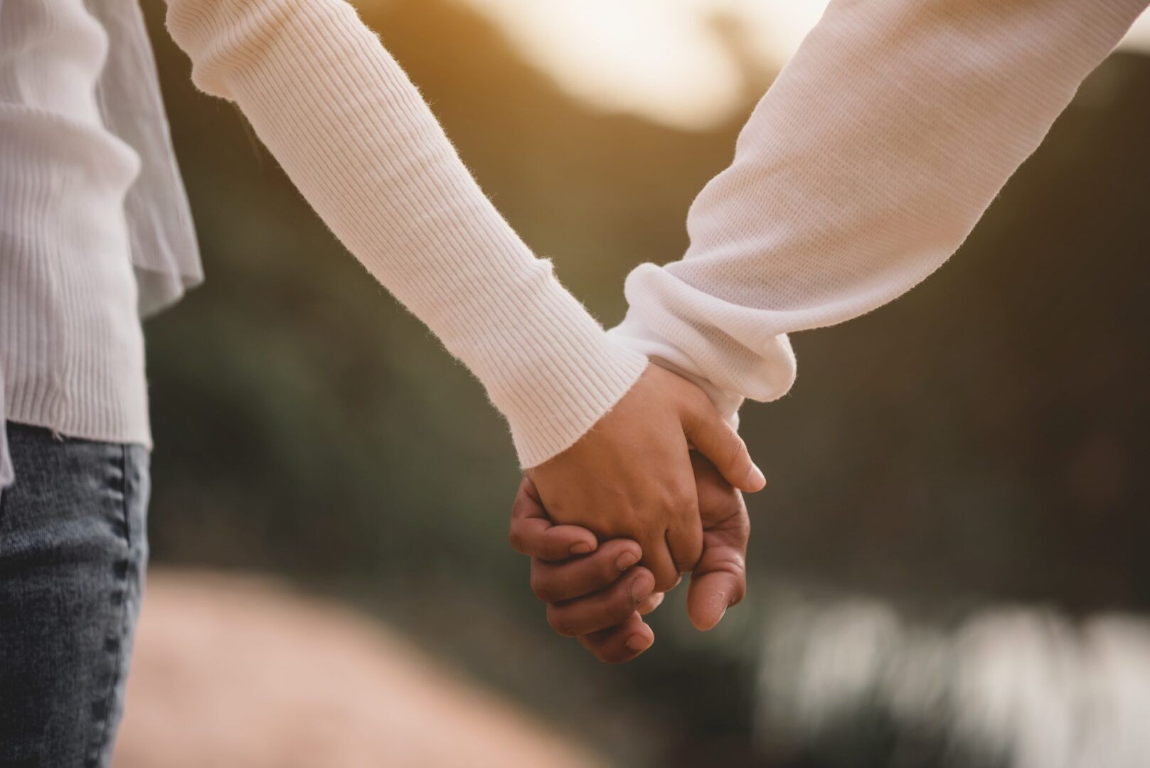 Two people wearing long-sleeved shirts are holding hands outdoors. The image is focused on their clasped hands, with a blurred natural background and warm lighting.