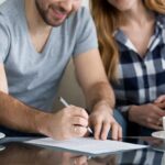 A man, smiling, signs a document on a glass table while a woman sits beside him holding his arm. Two cups and saucers are on the table. Both are casually dressed and partially visible.