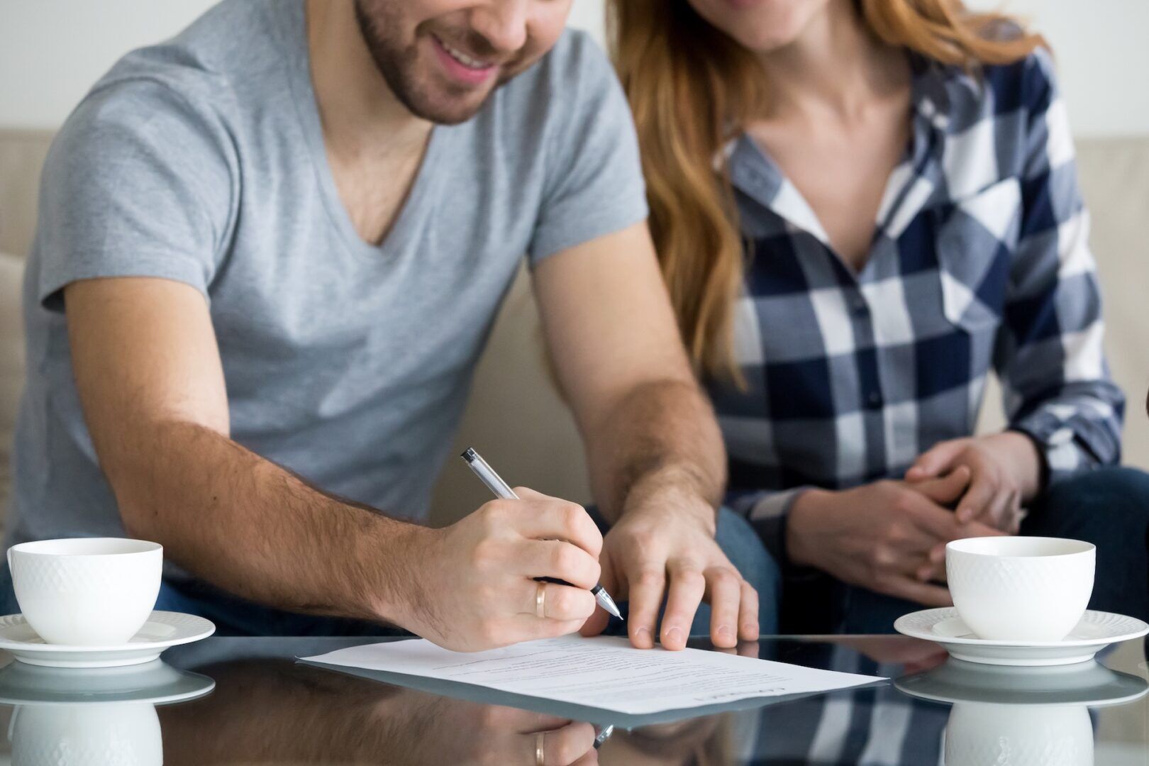 A man, smiling, signs a document on a glass table while a woman sits beside him holding his arm. Two cups and saucers are on the table. Both are casually dressed and partially visible.