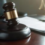 A judge’s gavel rests on a wooden desk next to a clipboard with papers and a pen. In the blurred background, a set of balance scales is visible, signifying justice and legal proceedings.