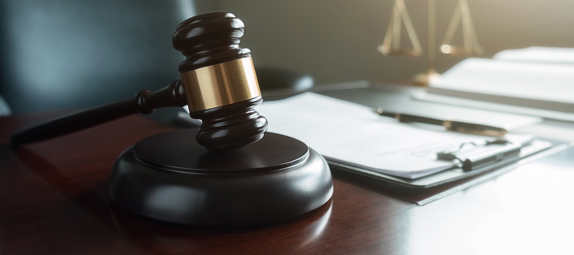 A judge’s gavel rests on a wooden desk next to a clipboard with papers and a pen. In the blurred background, a set of balance scales is visible, signifying justice and legal proceedings.