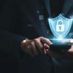 A person in a suit uses a smartphone with a glowing digital shield and padlock icon hovering above it, symbolizing cybersecurity and data protection.
