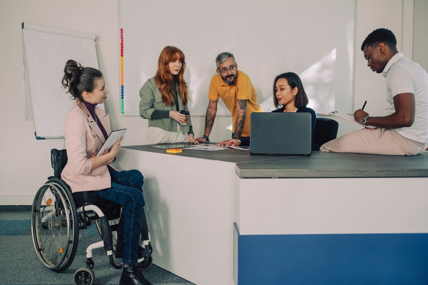 Five people having a discussion in an office; one woman uses a wheelchair, another works on a laptop, a man sits on the desk writing, and two others stand, one holding a coffee cup. A flipchart and whiteboard are in the background.